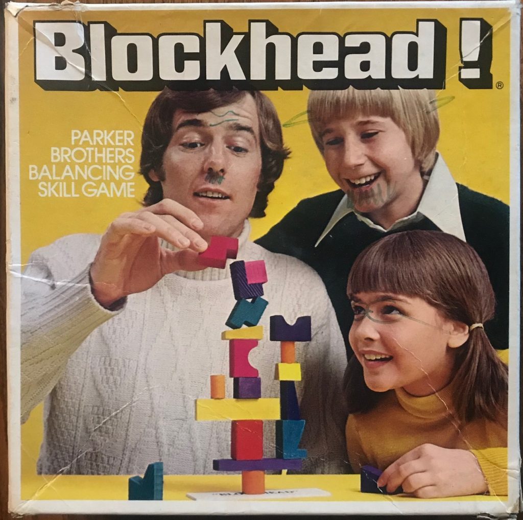 Two young children watch an older man try to delicately balance a block on top of a large structure